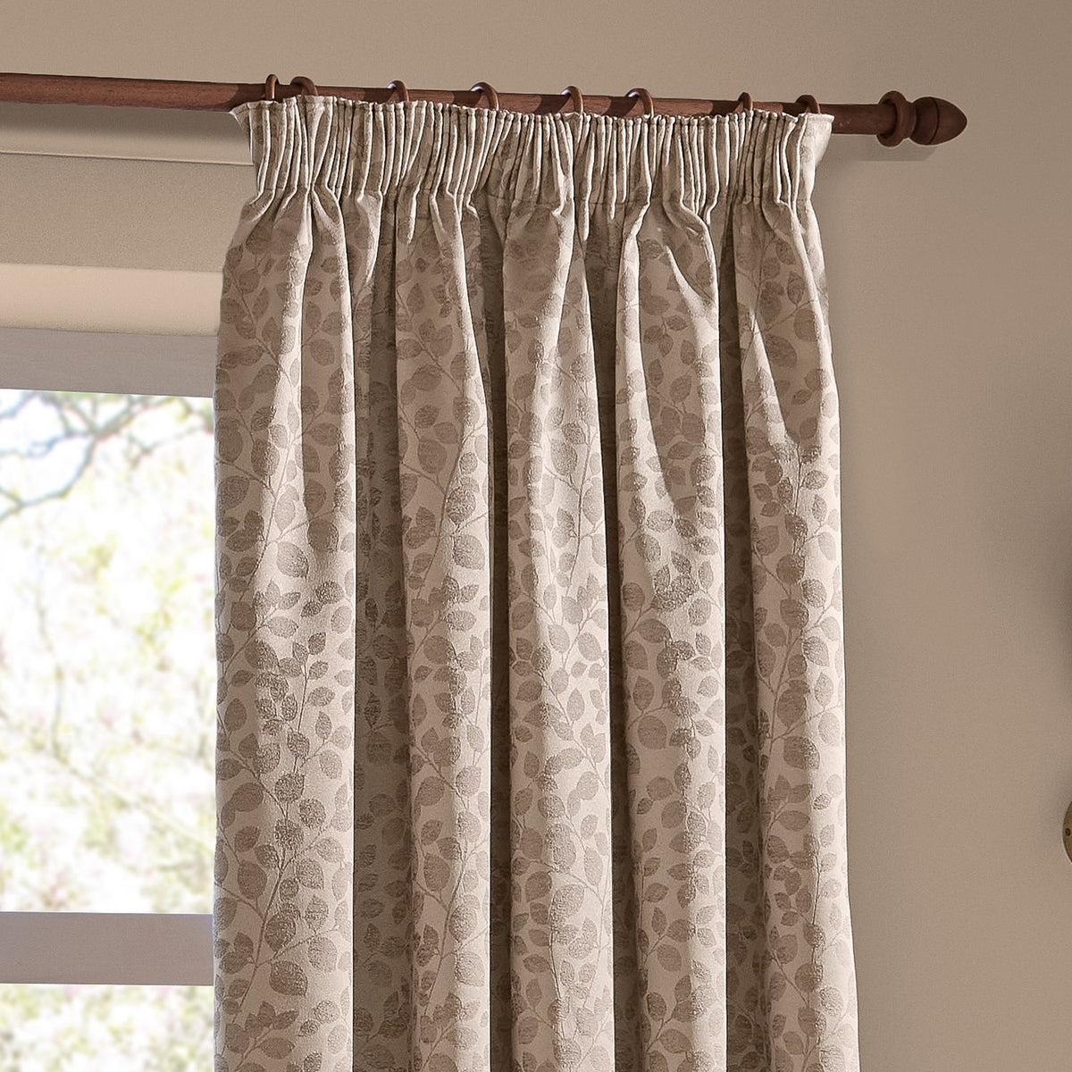 Grace Leaf Jacquard pencil pleat curtains in taupe, shown hanging on a wooden pole with a woven trailing leaf pattern in a living room window setting.
