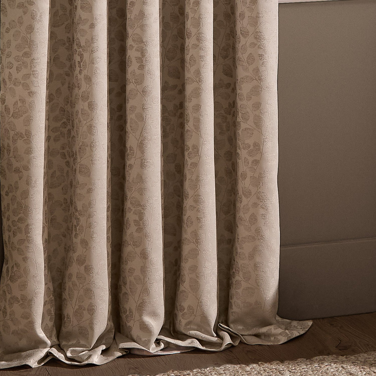 Grace Leaf Jacquard Pencil Pleat Curtains in taupe, pooling in soft folds with a woven leaf pattern, shown against a neutral wall and wood flooring.