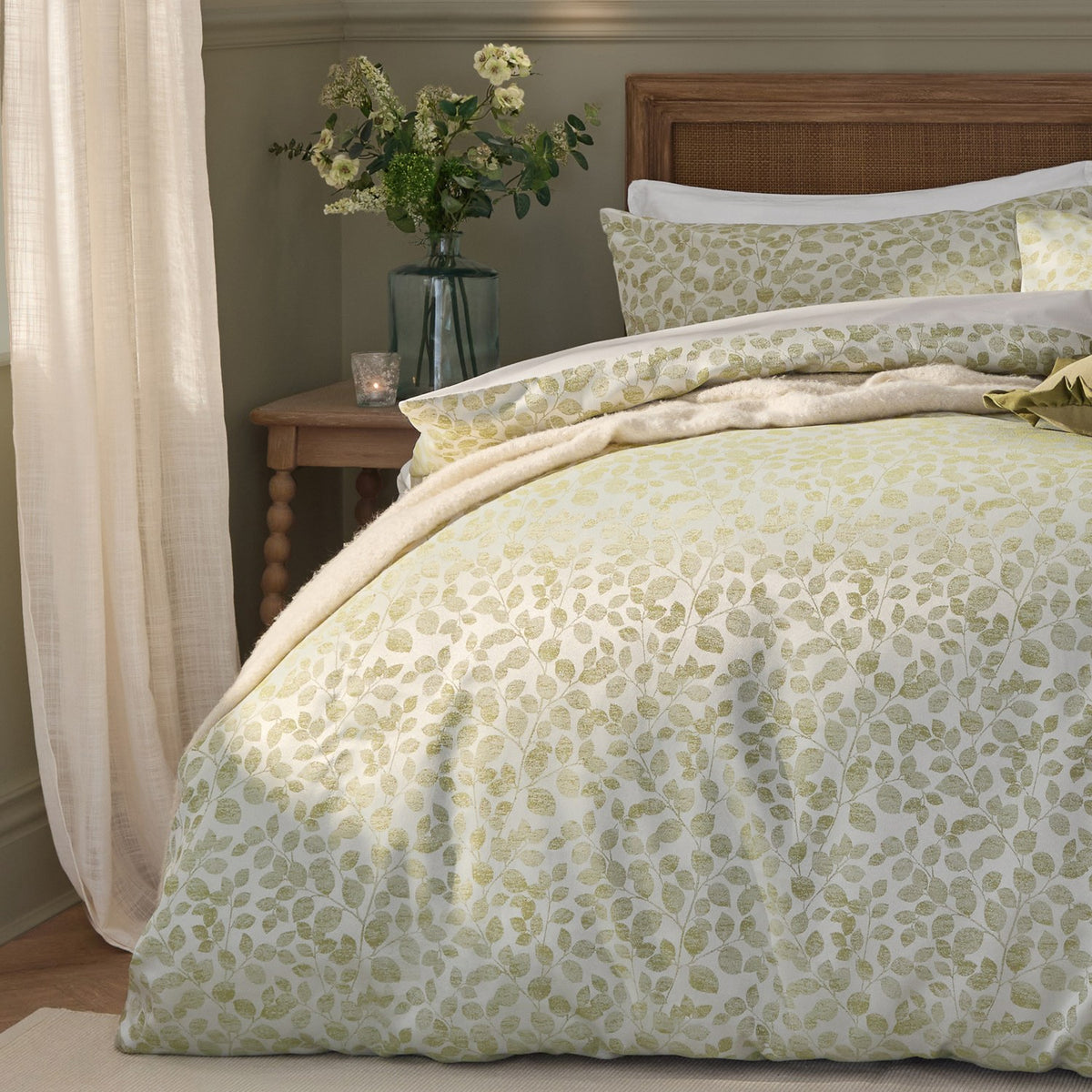 Grace Leaf Jacquard Floral Duvet Cover Set in Sage, shown on a bed with matching pillowcases, cream-green trailing leaf pattern, in a bedroom setting with wooden headboard and side table.