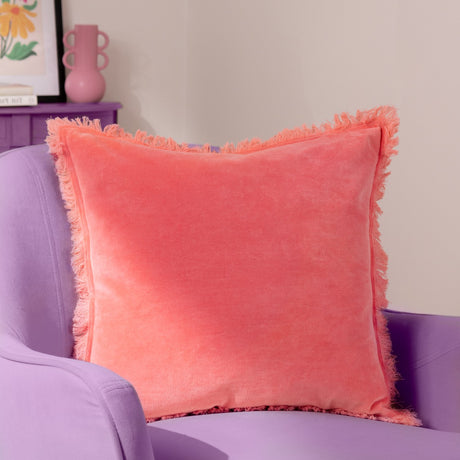 Gracie Velvet Cotton Cushion in coral, a square 45 x 45cm design with a soft brushed velvet face and fringed trim, shown on a lilac armchair in a living room setting.