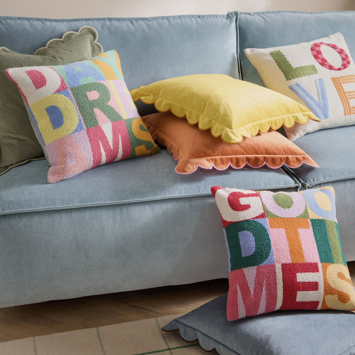Square tufted boucle cushion with "GOOD TIMES" in multicoloured block letters, shown on a light blue sofa with orange and yellow scalloped cushions nearby.