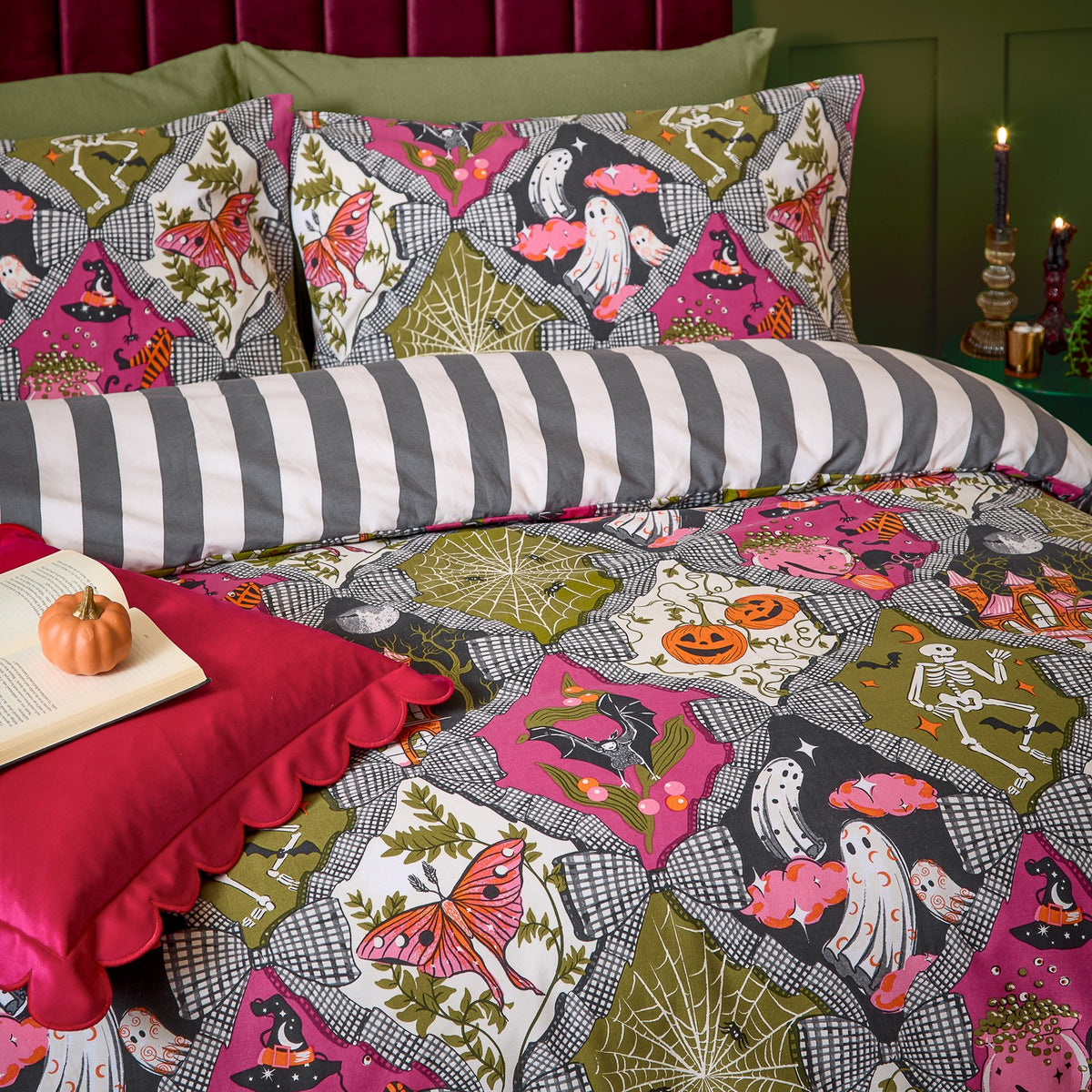 Hallowquin reversible duvet cover set on a bed with a Halloween ghosts, pumpkins, skeletons and cobwebs print in pink, olive and grey diamond panels, grey striped reverse and matching pillowcases.