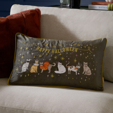 Rectangular grey velvet cushion with gold piping, showing embroidered Halloween cats in costumes and "Happy Halloween" text with gold stars, styled on a living room sofa.