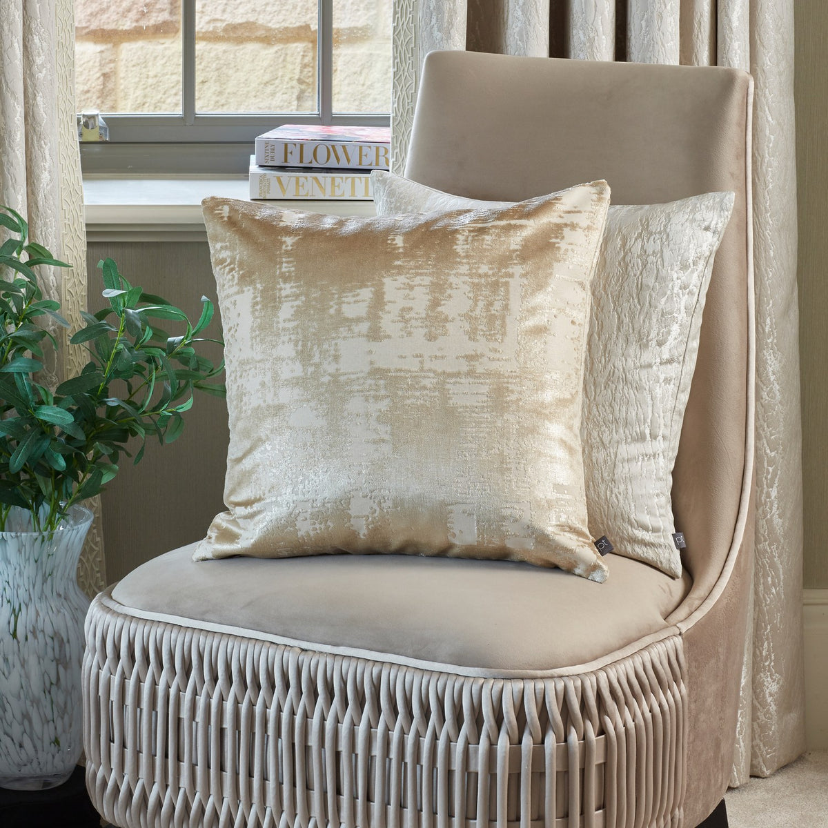 Hamlet Printed Polyester Cushion in Alabaster with a marbled metallic foil print on a cream ground, shown on a beige velvet chair in a living room setting.