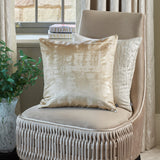 Hamlet Printed Polyester Cushion in Alabaster with a marbled metallic foil print on a cream ground, shown on a beige velvet chair in a living room setting.