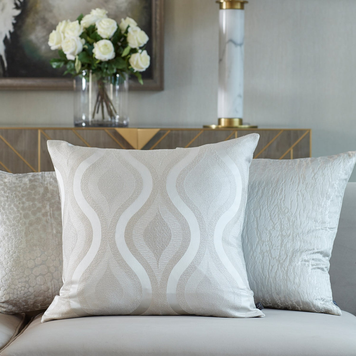 Hamlet Printed Polyester Cushion in Alabaster with a wavy metallic pattern on a cream ground, shown on a sofa alongside textured accent cushions in a living room setting.
