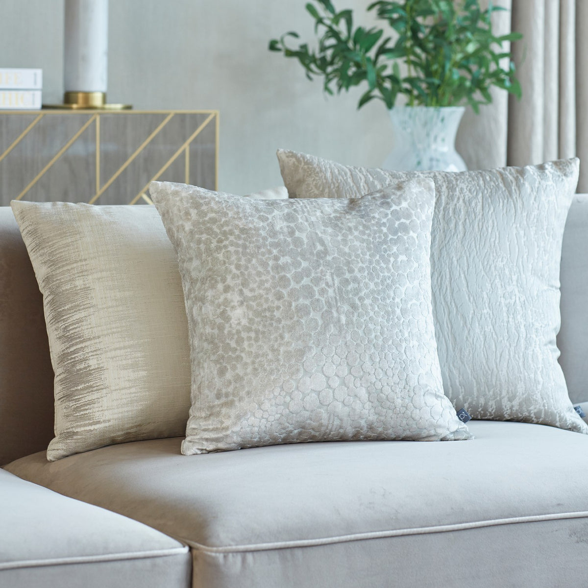 Hamlet Printed Polyester Cushion in Alabaster, a square cream cushion with a marbled metallic foil pattern, styled with two neutral cushions on a beige sofa.
