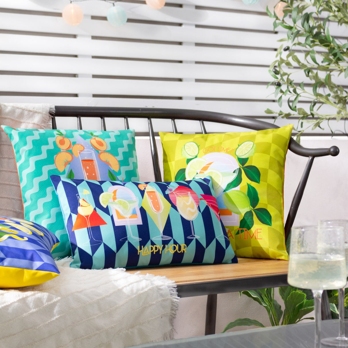 Happy Hour rectangular cushion in navy and pale aqua geometric print with cocktail glasses and yellow slogan text, shown on an outdoor bench with other patterned cushions.