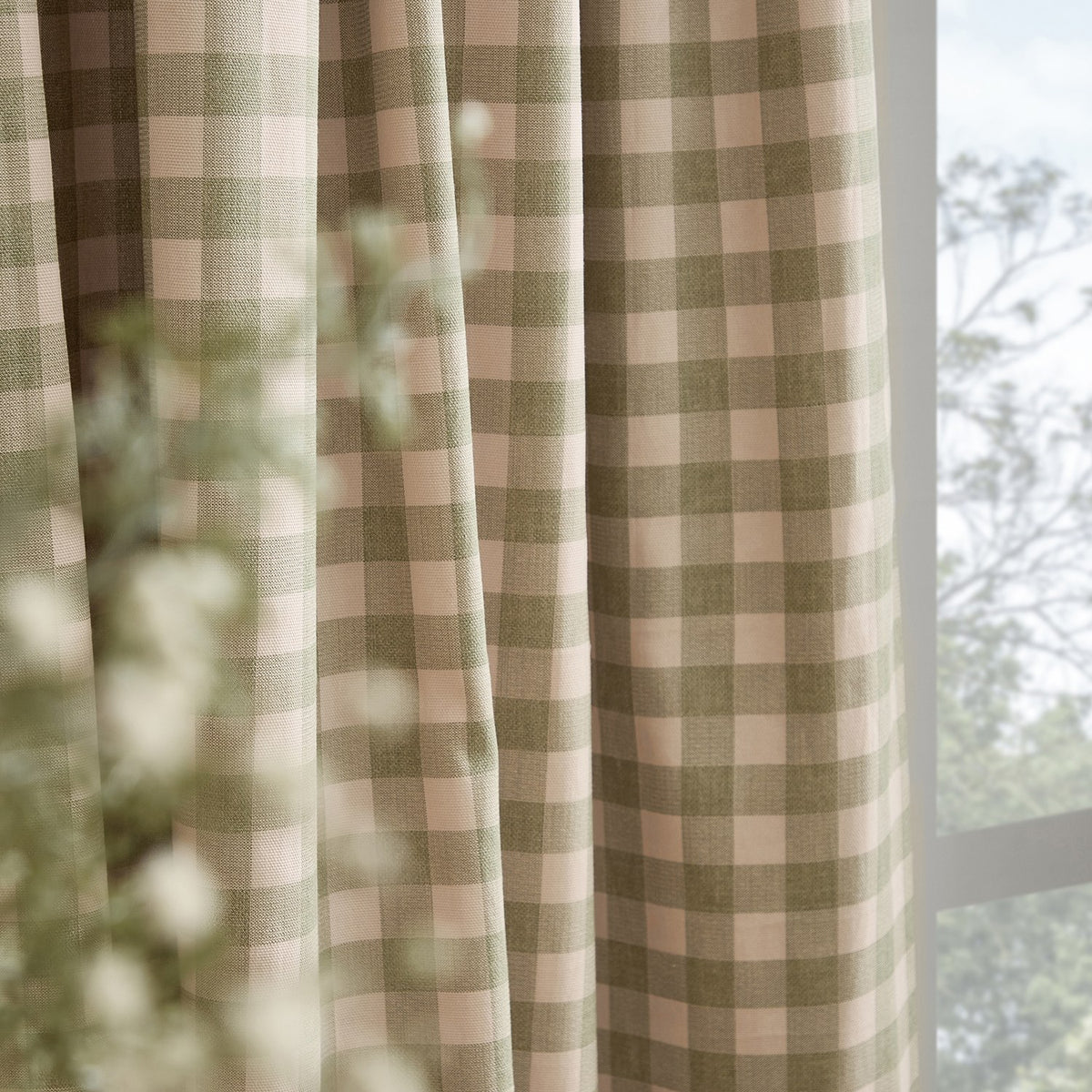 Harriet Gingham Cotton Pencil Pleat Curtains in pink and green check fabric, shown draped in folds beside a window with softly blurred foliage.