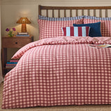 Harriet Gingham Cotton Check Duvet Cover Set in Burgundy/Blossom on a bed with striped and blue cushions, wooden headboard, and bedside table with lamp.