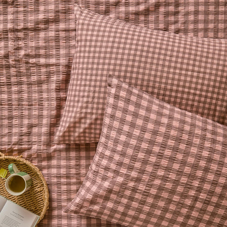 Harriet Gingham Cotton Pillowcase Pair in rose and chocolate, shown on a matching gingham seersucker duvet cover with a woven tray, mug, and book nearby.