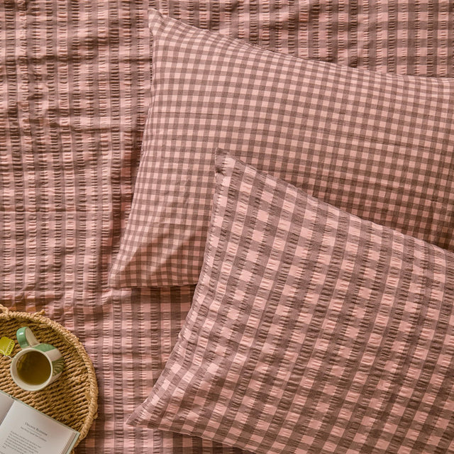 Harriet Gingham Cotton Pillowcase Pair in rose and chocolate, shown on a matching gingham seersucker duvet cover with a woven tray, mug, and book nearby.