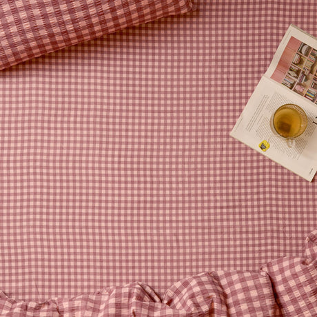 Harriet fitted sheet in burgundy and blossom gingham check cotton with a ruffled edge, shown on a bed beside a matching pillowcase, open magazine, and cup of tea.