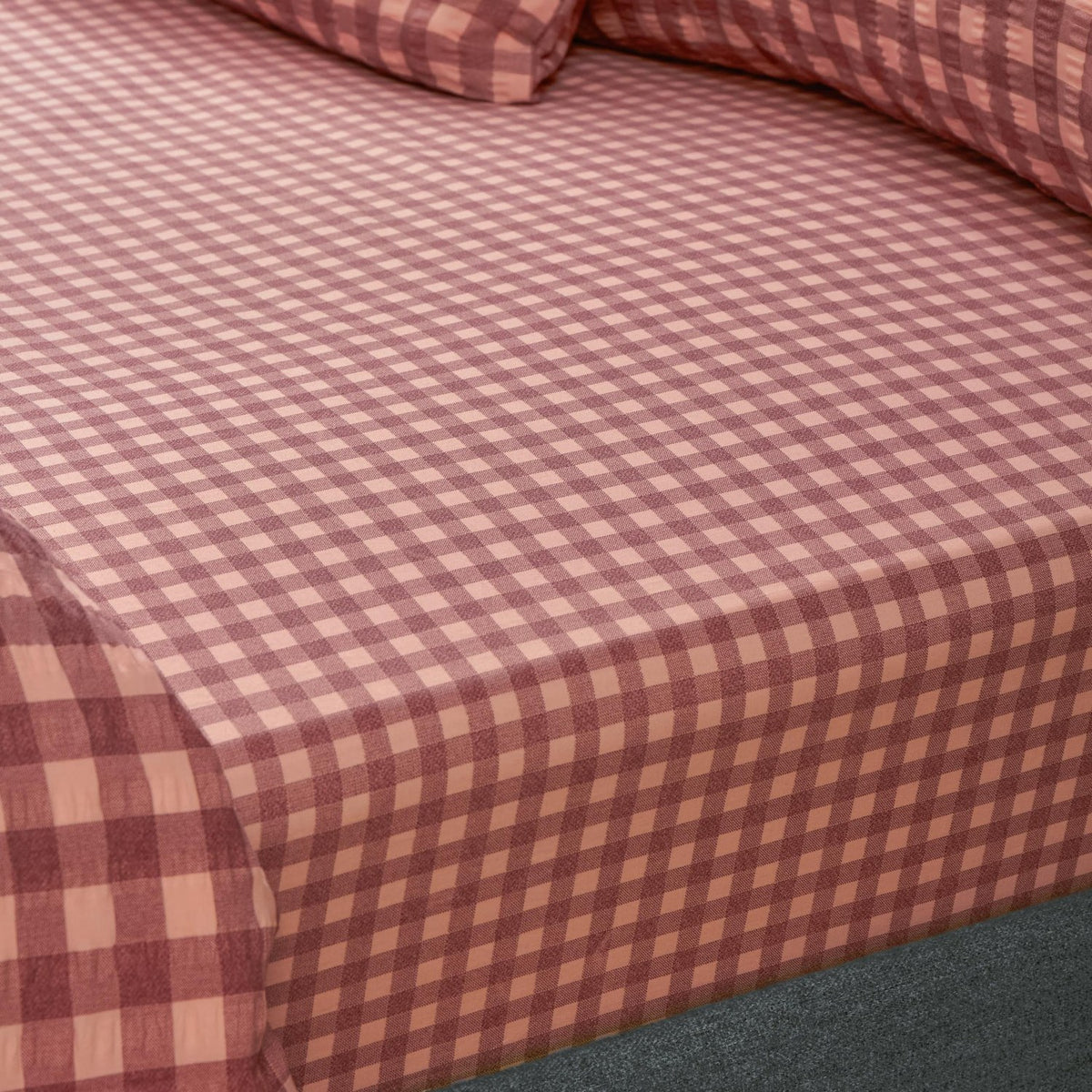 Corner view of the Harriet fitted sheet in burgundy and blossom gingham check cotton, showing the elasticated edge fitted over a mattress with matching pillowcase behind.