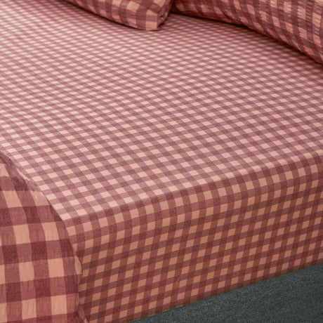 Corner view of the Harriet fitted sheet in burgundy and blossom gingham check cotton, showing the elasticated edge fitted over a mattress with matching pillowcase behind.