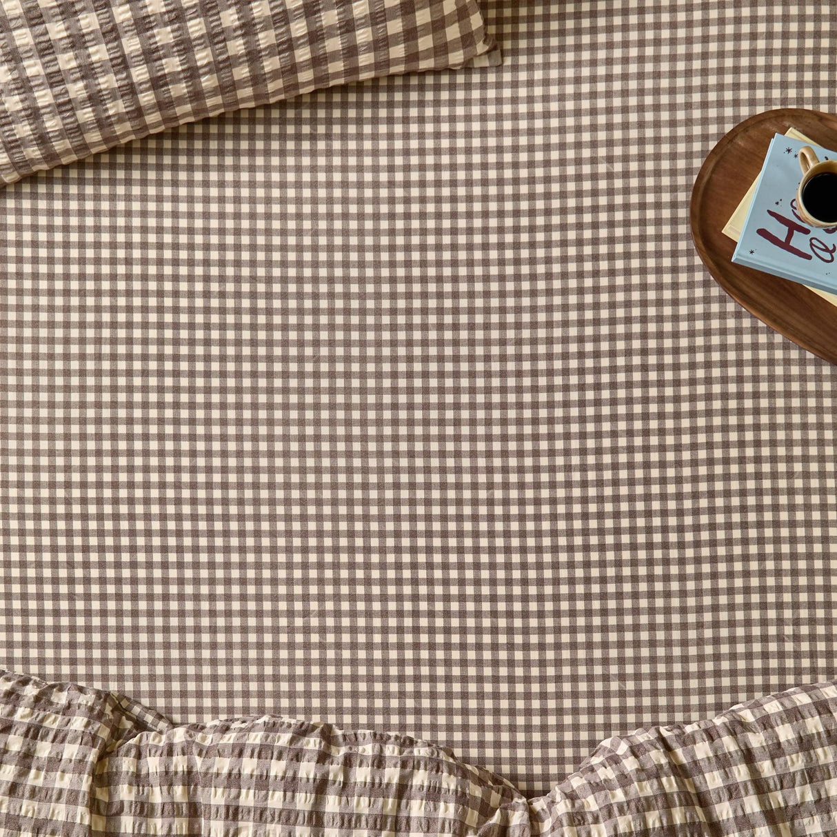 Harriet Gingham Check Cotton Fitted Sheet in buttermilk and chocolate, shown on a bed with matching pillowcase and duvet, styled beside a wooden tray with coffee and books.