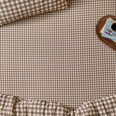 Harriet Gingham Check Cotton Fitted Sheet in buttermilk and chocolate, shown on a bed with matching pillowcase and duvet, styled beside a wooden tray with coffee and books.