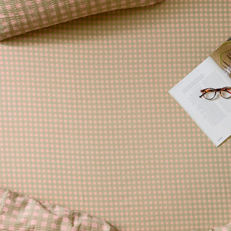 Harriet Gingham Check Cotton Fitted Sheet in pink and green, shown on a bed with matching seersucker cushions, an open magazine and glasses resting on top.