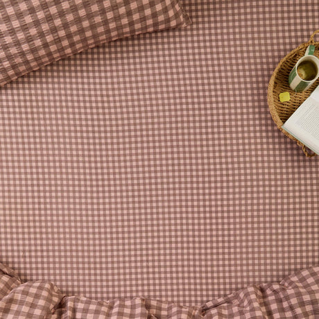 Harriet Gingham Check Cotton Fitted Sheet in rose and chocolate on a bed, shown with a matching pillowcase, ruffled edge, and a bedside tray with book and mug.