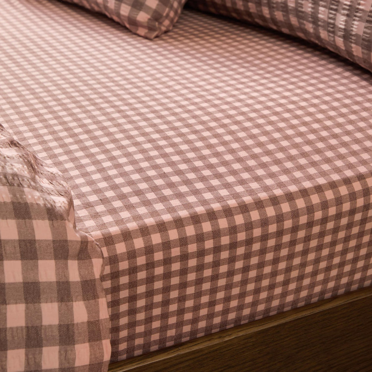 Harriet Gingham Check Cotton Fitted Sheet in rose and chocolate, showing the elasticated corner fitted over a mattress with matching pillowcases in the background.