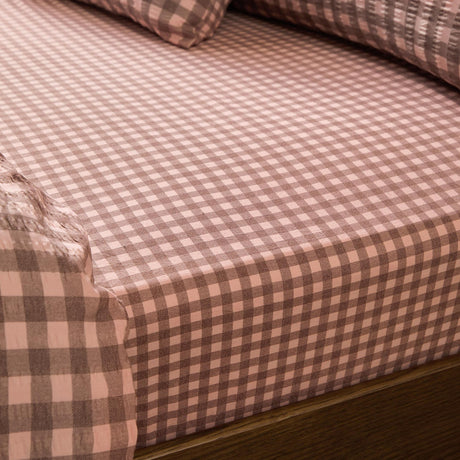 Harriet Gingham Check Cotton Fitted Sheet in rose and chocolate, showing the elasticated corner fitted over a mattress with matching pillowcases in the background.