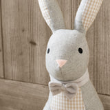 Close-up of the Harvey Hare Check Cotton Door Stop showing grey herringbone face and ears, beige gingham lining, pink nose, embroidered eyes and a grey bow tie against a wooden background.
