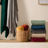 Harrow Plain Polyester Throw in grey, folded flannel fleece shown on a shelf beside stacked throws in green, blue, beige and red, with two rolled throws in a woven basket.