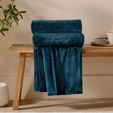 Harrow Plain Polyester Throw in Midnight, a folded flannel fleece throw in deep teal-blue draped over a wooden bench in a living room setting.