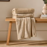 Harrow Plain Polyester Throw in natural cream, folded on a wooden bench with part draped down, showing its smooth flannel fleece texture in a living room setting.