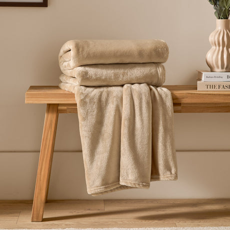 Harrow Plain Polyester Throw in natural cream, folded on a wooden bench with part draped down, showing its smooth flannel fleece texture in a living room setting.