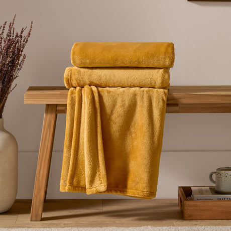 Harrow Plain Polyester Throw in ochre yellow, folded and draped over a wooden bench in a living room setting, showing its smooth flannel fleece texture.