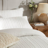 Harrison Stripe Tufted Polyester Duvet Cover Set in white on a bed with matching pillowcases and a cushion, styled in a bedroom beside a wooden nightstand with a lamp and vase of flowers.