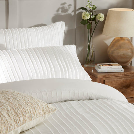 Harrison Stripe Tufted Polyester Duvet Cover Set in white on a bed with matching pillowcases and a cushion, styled in a bedroom beside a wooden nightstand with a lamp and vase of flowers.