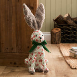 Hattie polyester door stop shaped like a sitting hare, with brown bouclé ears, cream floral-print body, and a green ribbed bow, shown on a wooden floor near a rustic hallway setting.