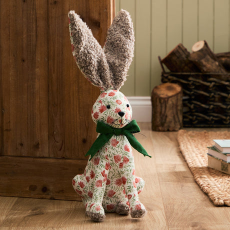 Hattie polyester door stop shaped like a sitting hare, with brown bouclé ears, cream floral-print body, and a green ribbed bow, shown on a wooden floor near a rustic hallway setting.