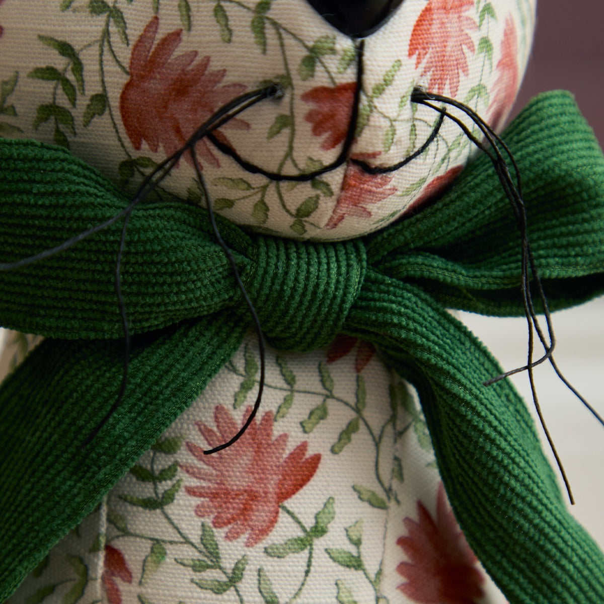 Close-up of the Hattie Polyester Door Stop, a hare-shaped design with a cream floral fabric face, black nose and eyes, and a hand-tied green ribbed bow at the neck.