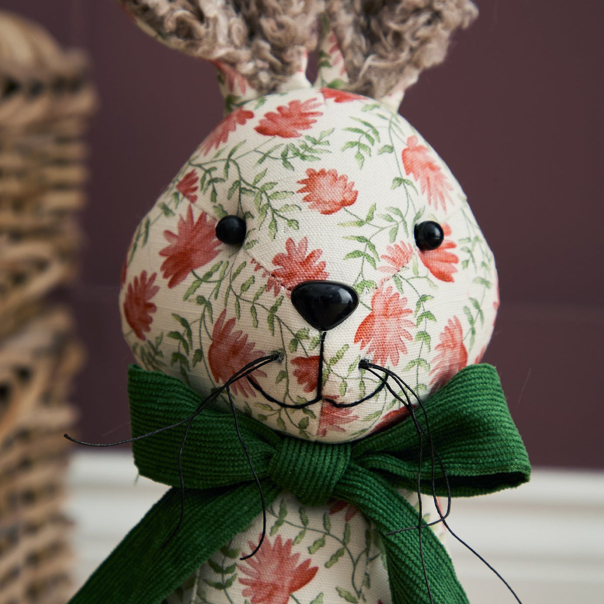 Hattie Polyester Door Stop hare-shaped design, close-up of face with bouclé ears, floral cream fabric, black button eyes and nose, green ribbed bow at neck.
