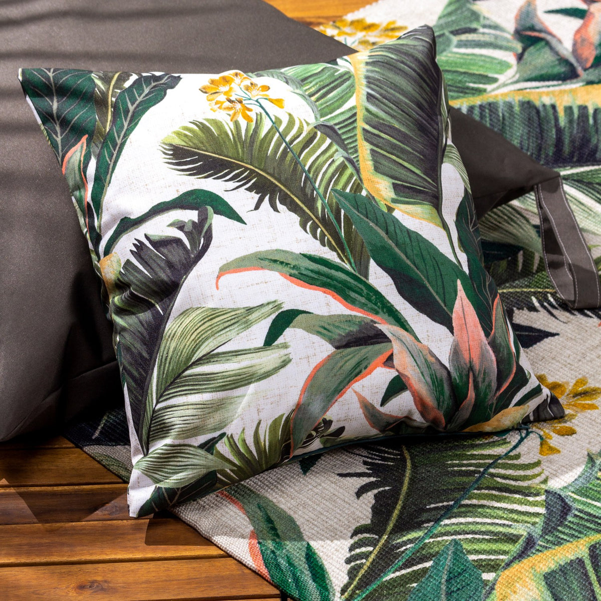 Hawaii Jungle Polyester Cushion in Multi, a square cushion with a pale ground and green tropical leaf print, amber-gold flowers and coral-pink stems, shown on a sofa in a living room setting.
