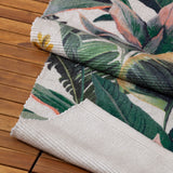 Hawaii Floral Polyester Rug in Green, folded to show flatweave texture with jungle leaf print in green, pink and yellow on a white base, plain woven reverse, shown on wooden decking.