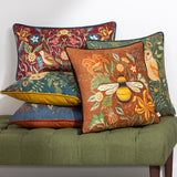 Square ginger chenille cushion with a hand-painted bumblebee and woodland floral design, shown among patterned cushions on a green upholstered bench.