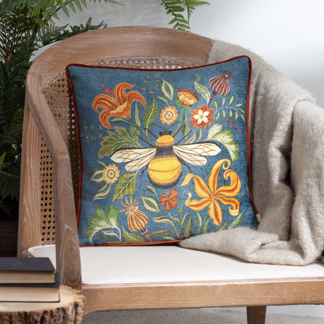 Hawthorn printed velvet chenille cushion in petrol blue with bumblebee and floral design, rust piping, styled on a rattan-back chair with a woven throw.