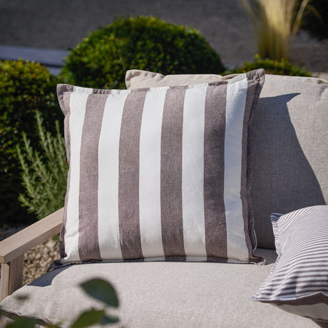 Hayle Reversible Cotton Cushion in Chocolate, 50x50cm square with wide brown and white stripes, shown on an outdoor sofa amid garden greenery.