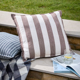 Hayle Reversible Cotton Cushion in Chocolate, 50x50cm square with wide brown and white stripes, shown outdoors on a stone ledge beside a striped blue cushion.