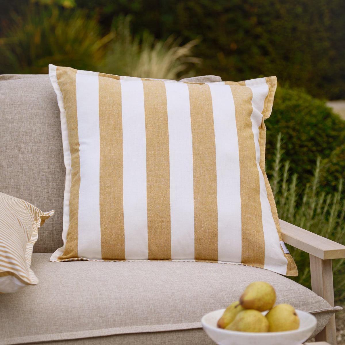 Hayle Reversible Cotton Cushion in honey and white wide stripes, 50 x 50cm, shown on a sofa in an outdoor garden setting.