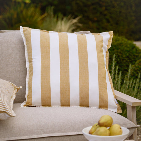 Hayle Reversible Cotton Cushion in honey and white wide stripes, 50 x 50cm, shown on a sofa in an outdoor garden setting.