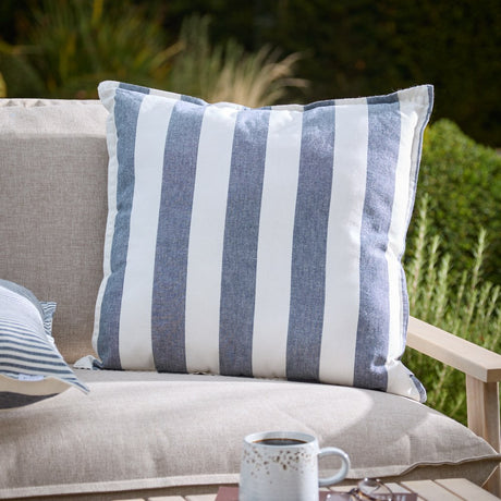 Hayle Reversible Cotton Cushion in navy, square with wide navy-and-white stripes, shown on a beige outdoor sofa in a garden setting.