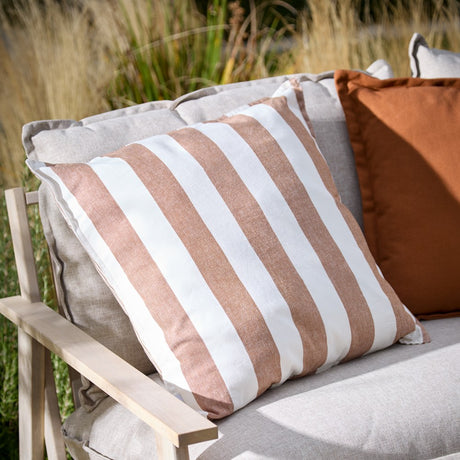 Hayle Reversible Cotton Cushion in pecan brown and white wide stripes, styled on an outdoor sofa with a rust cushion, set against a garden backdrop.