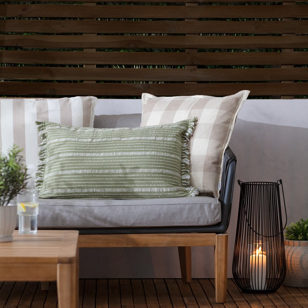 Outdoor lounge setting with a wooden-framed sofa styled with beige check and green striped cushions, evoking the Hayle Reversible Cotton Seat Pad's natural palette, plus a lit lantern and side table.