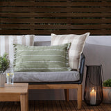 Outdoor lounge setting with a wooden-framed sofa styled with beige check and green striped cushions, evoking the Hayle Reversible Cotton Seat Pad's natural palette, plus a lit lantern and side table.