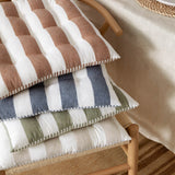 Hayle reversible cotton seat pads in striped rust, blue, green, and natural stripes, stacked on a wooden chair beside a table setting.